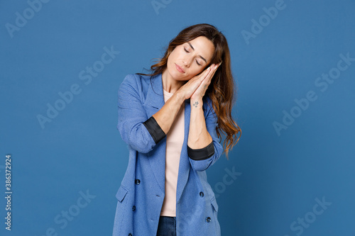 Relaxed tired beautiful attractive young brunette woman 20s wearing basic jacket sleep with folded hands under cheek keeping eyes closed isolated on bright blue colour background, studio portrait.