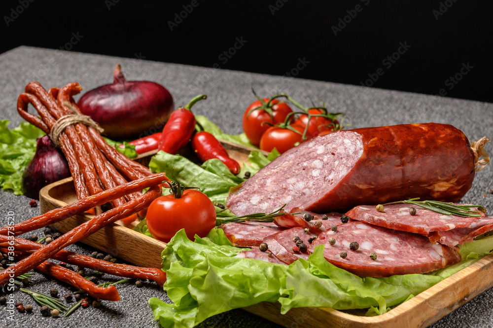 The slices sausage served on lettuce leaves in a wooden plate. Also thin long sticks of smoked sausage tied with string. Chili pepper, tomatoes on a sprig, onion and garlic. Close-up photo