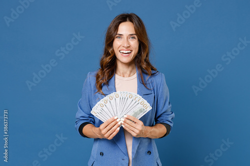 Fotografie Laughing funny young brunette woman 20s wearing basic jacket standing holding in hand fan of cash money in dollar banknotes looking camera isolated on bright blue colour background studio portrait
