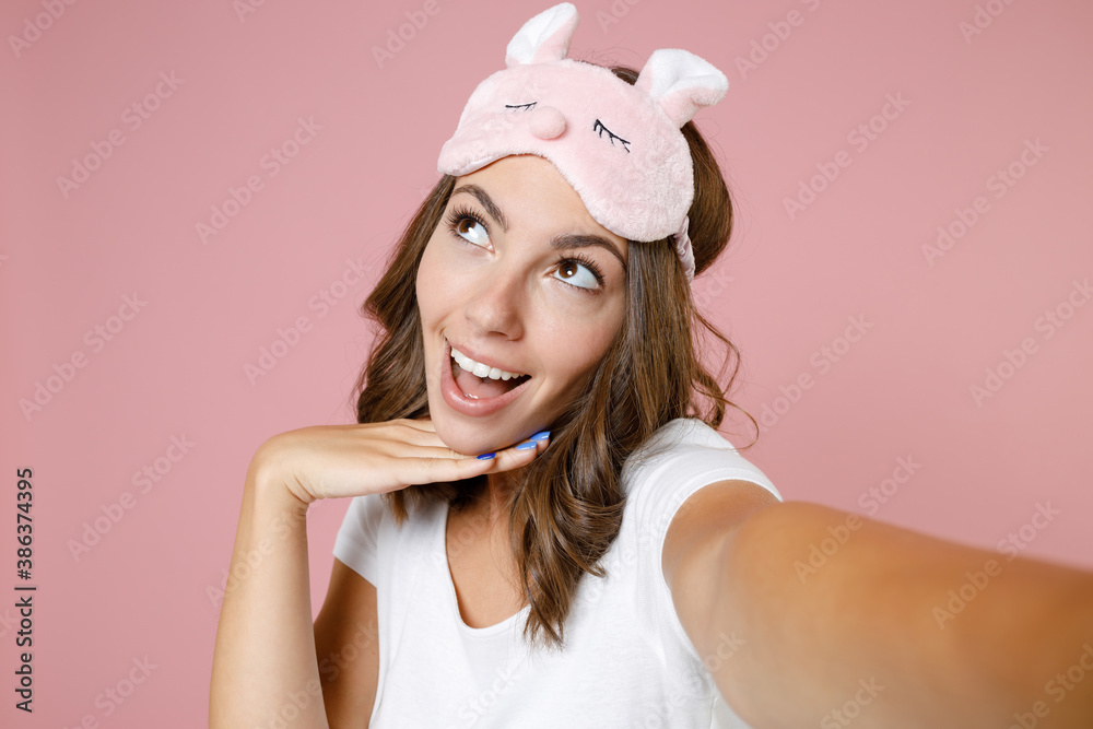 Close up of excited young woman in pajamas home wear sleep mask doing selfie shot on mobile phone put hand prop up on chin resting at home isolated on pink background studio. Relax good mood concept.