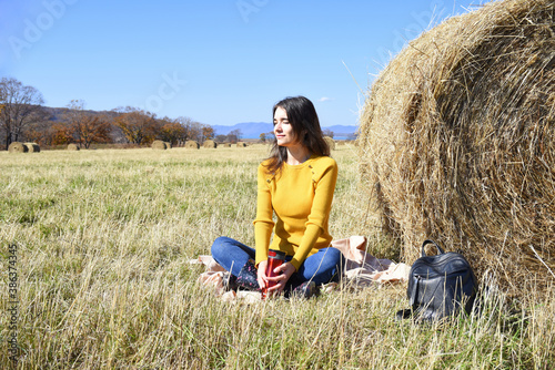 Young Russian brunette woman in yellow sweater and rubber boots sitting on plaid next to backpack near bale of hay in field. she holding thermos with tea,  from the sun and smiling.  © Tatiana