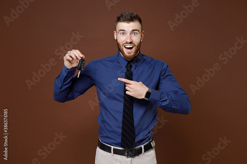 Excited surprised bearded young business man wearing blue shirt tie pointing index finger on car keys isolated on brown colour background studio portrait. Achievement career wealth business concept.