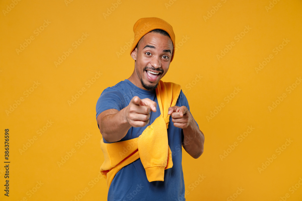 Surprised cheerful excited young african american man 20s wearing basic casual blue t-shirt hat standing pointing index fingers on camera isolated on bright yellow colour background, studio portrait.