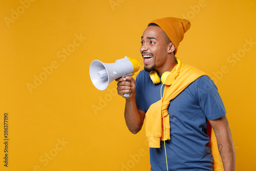 Excited cheerful funny young african american man 20s wearing basic casual blue t-shirt hat standing screaming in megaphone looking aside isolated on bright yellow colour background, studio portrait.