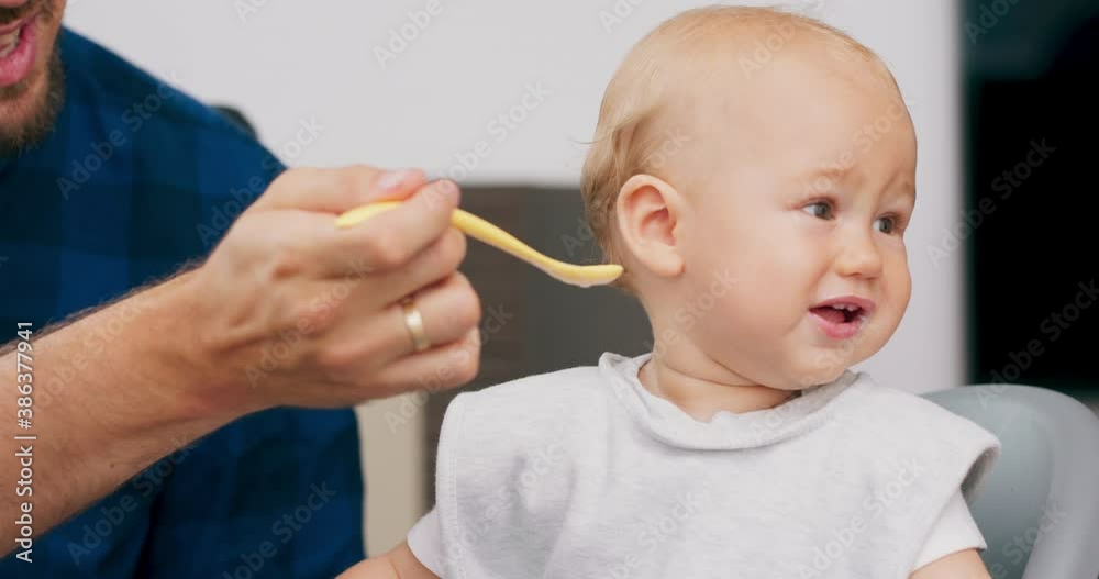 Closeup cute baby with bib, in high baby chair. Young bearded father feeds baby with spoon. Baby is spinning in the chair, grimaces. Slow motion Closeup cute baby with bib, in high baby chair. Young bearded father feeds baby with spoon. Baby is spinning in the chair, grimaces. Slow motion