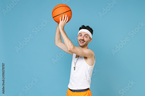 Excited young sporty fitness man basketball player with thin skinny body sportsman in headband shirt shorts whistle playing hold ball isolated on blue background. Workout gym sport motivation concept.