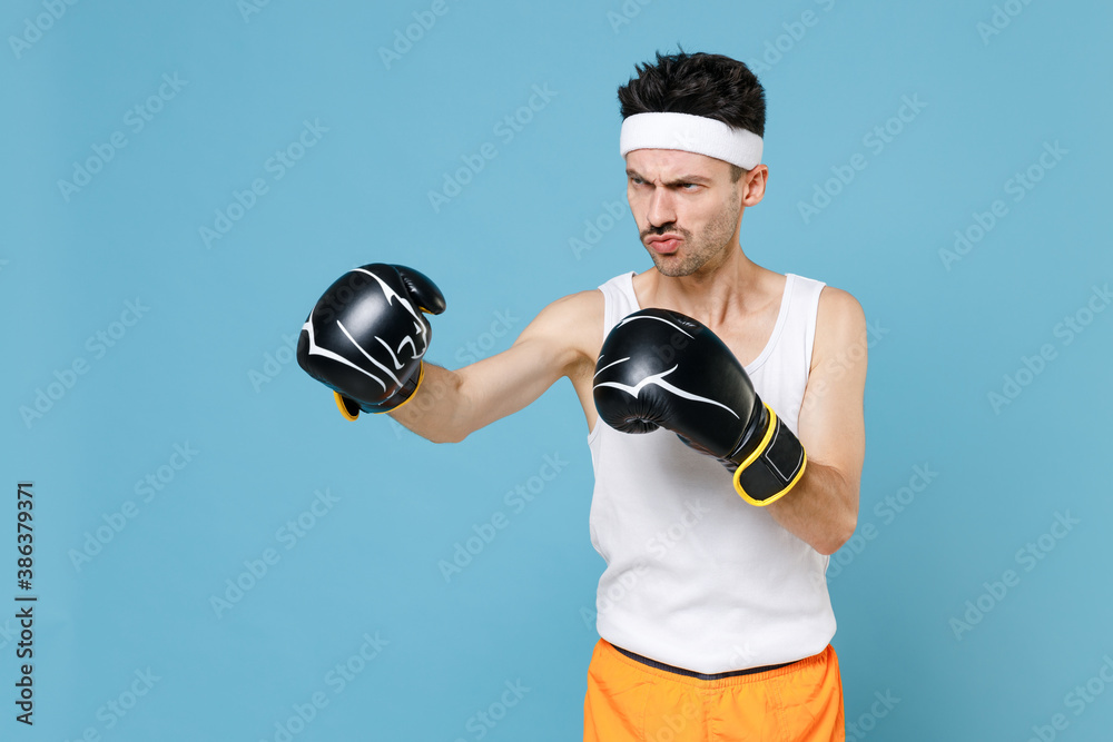 Confused young strong sporty fitness man boxer with skinny body sportsman in headband shirt shorts doing boxing exercises in gloves isolated on blue background. Workout gym sport motivation concept. Confused young strong sporty fitness man boxer with skinny body sportsman in headband shirt shorts doing boxing exercises in gloves isolated on blue background. Workout gym sport motivation concept.