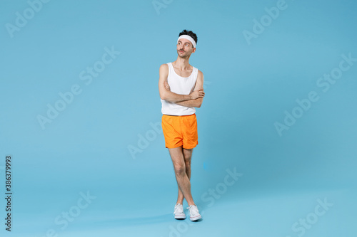 Full length portrait of young sporty fitness man with skinny body sportsman in white headband shirt shorts holding hands crossed isolated on blue background. Workout gym sport motivation concept.
