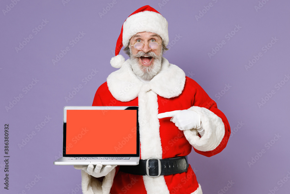 Excited Santa Claus man in Christmas hat red coat pointing index finger on laptop pc computer with blank empty screen isolated on violet background. Happy New Year celebration merry holiday concept. Excited Santa Claus man in Christmas hat red coat pointing index finger on laptop pc computer with blank empty screen isolated on violet background. Happy New Year celebration merry holiday concept.
