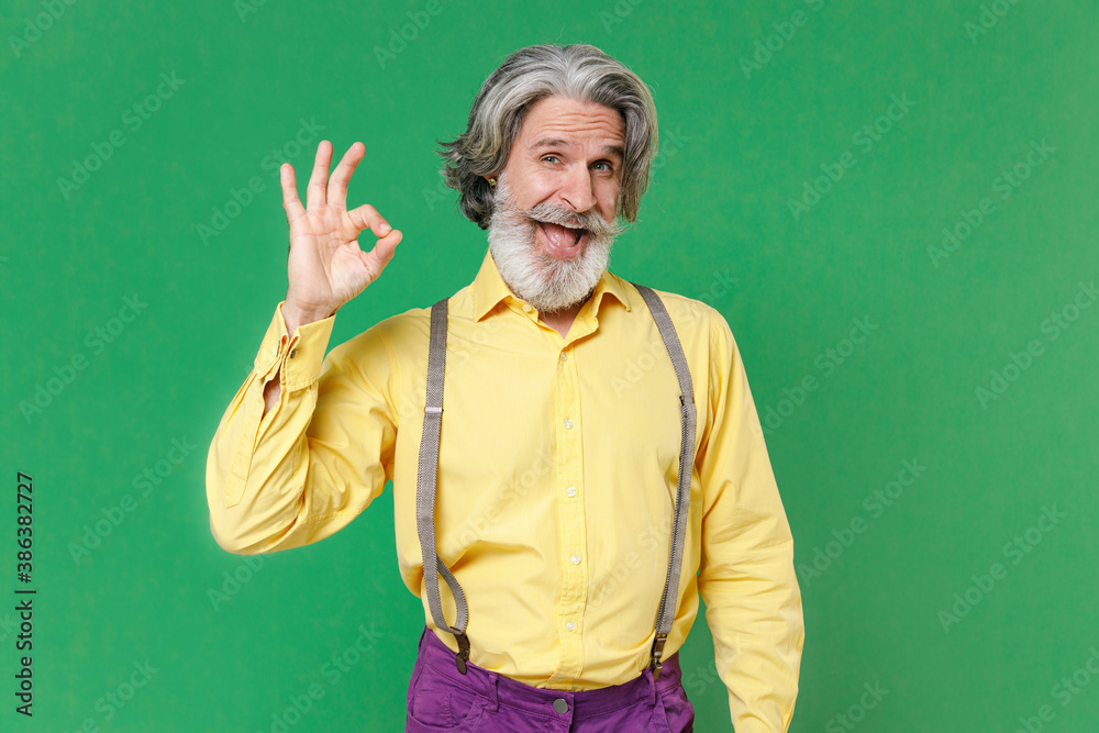 Excited cheerful elderly gray-haired mustache bearded man wearing casual yellow shirt suspenders standing showing Ok gesture looking camera isolated on bright green colour background, studio portrait.