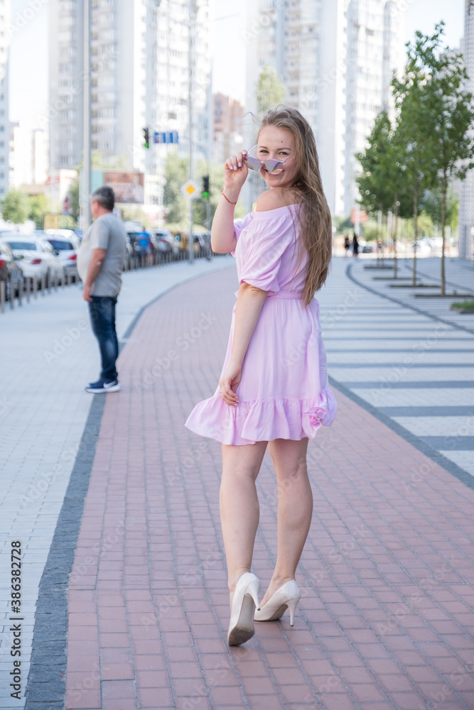 Cute cheerful woman in pink dress stand on a city street and smile. Summer day portrait.