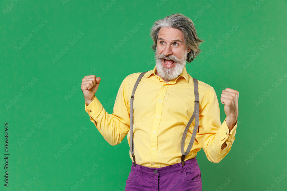 Excited joyful elderly gray-haired mustache bearded man in basic yellow shirt suspenders clenching fists doing winner gesture looking camera isolated on bright green colour background studio portrait.
