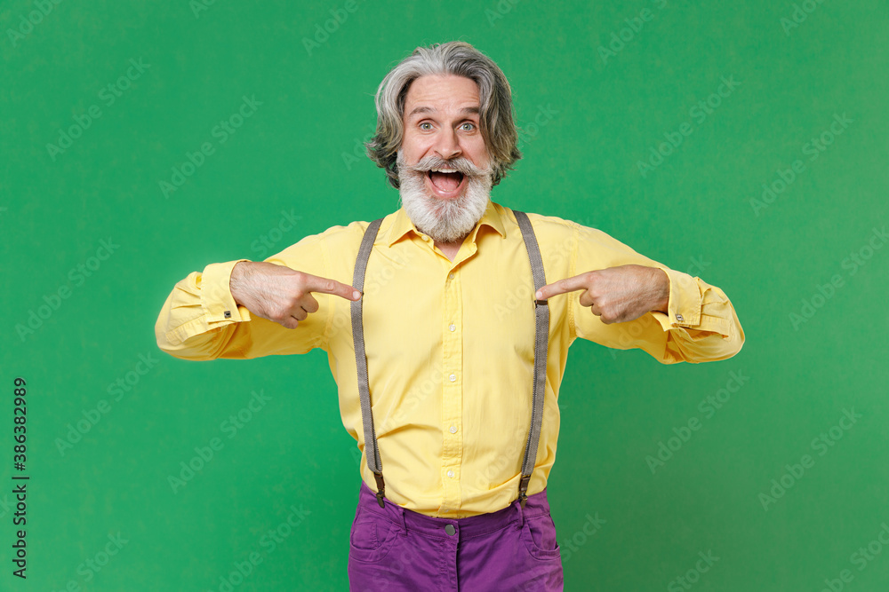 Surprised elderly gray-haired mustache bearded man wearing basic yellow shirt suspenders pointing index fingers on himself looking camera isolated on bright green colour background studio portrait.