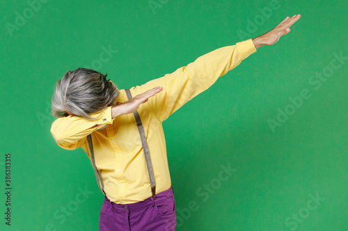 Elderly gray-haired mustache bearded man wearing casual yellow shirt suspenders doing dab hip hop dance hands gesture, youth sign hiding and covering face isolated on green background studio portrait.