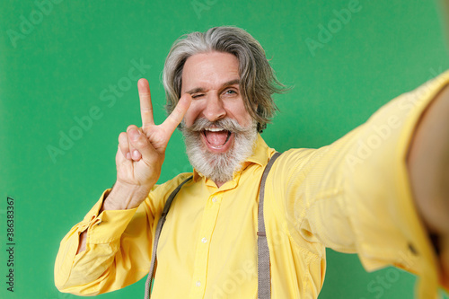Close up of blinking elderly gray-haired mustache bearded man wearing basic yellow shirt suspenders doing selfie shot on mobile phone showing victory sign isolated on green background studio portrait. © ViDi Studio