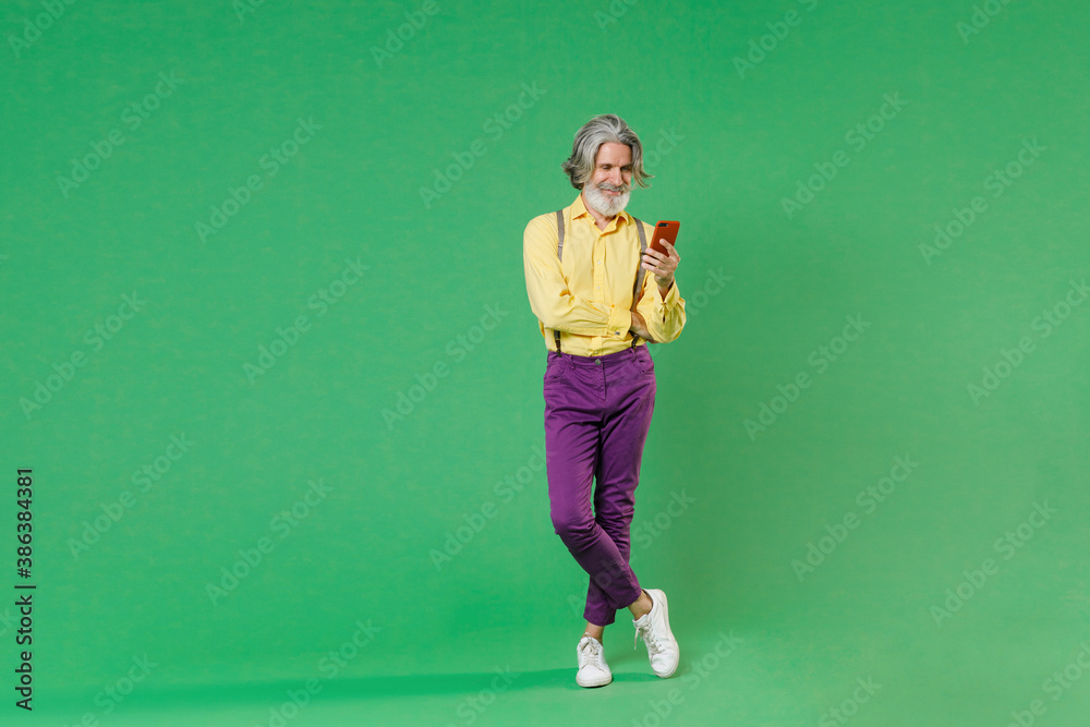 Full length of smiling elderly gray-haired mustache bearded man in basic yellow shirt suspenders using mobile cell phone typing sms message isolated on bright green colour background, studio portrait.