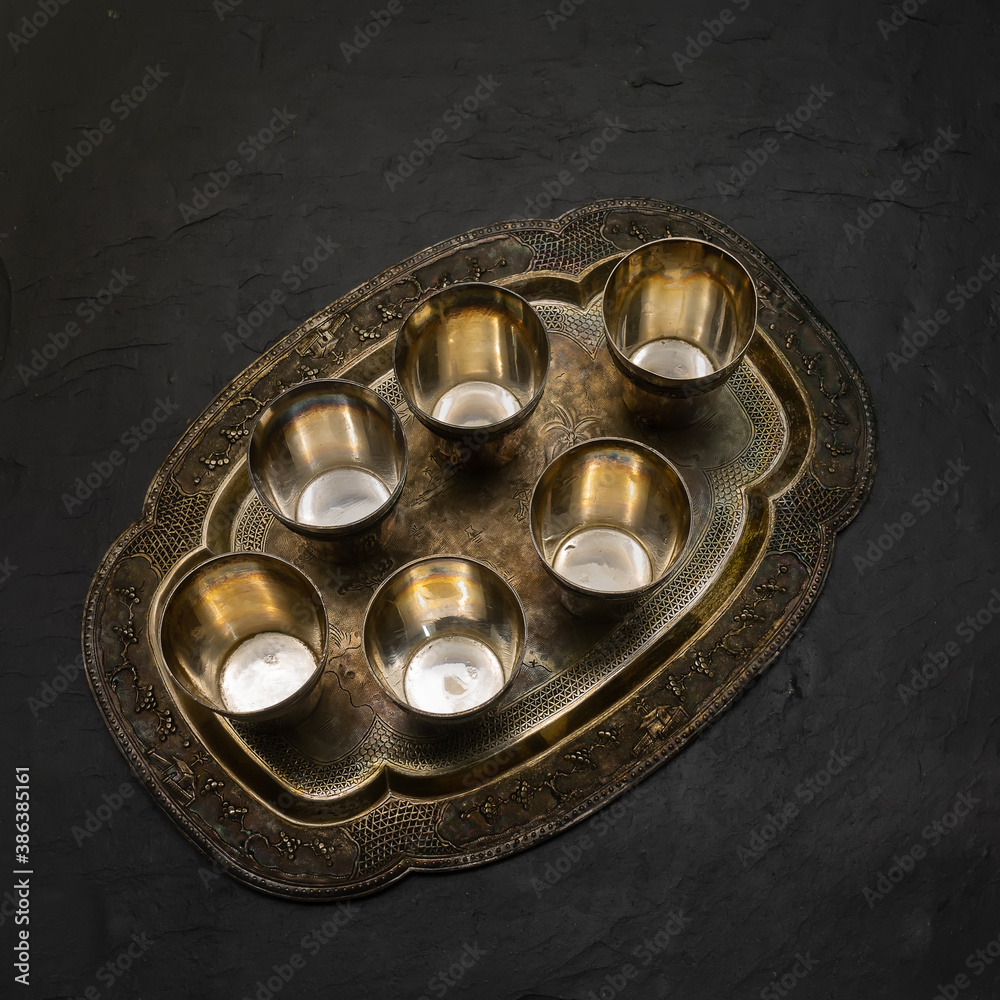 Set of antique silver items. Six small glasses on a curly tray. Decoration - ornaments on Asian themes. Covered with patina. 20th century. The background is black stone tiles. View from above. Set of antique silver items. Six small glasses on a curly tray. Decoration - ornaments on Asian themes. Covered with patina. 20th century. The background is black stone tiles. View from above.