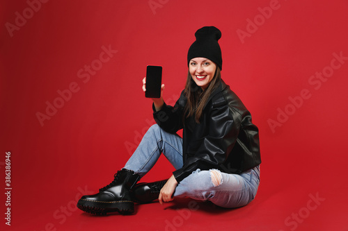 Full length of smiling funny young brunette woman in black leather jacket white t-shirt hat sitting hold mobile phone with blank empty screen looking camera isolated on red background studio portrait.