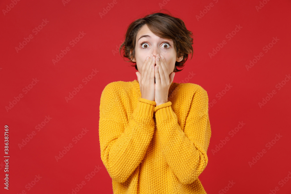 Shocked amazed surprised excited young brunette woman 20s wearing basic casual yellow sweater stand covering mouth with hands looking camera isolated on bright red colour background studio portrait. - obrazy, fototapety, plakaty 