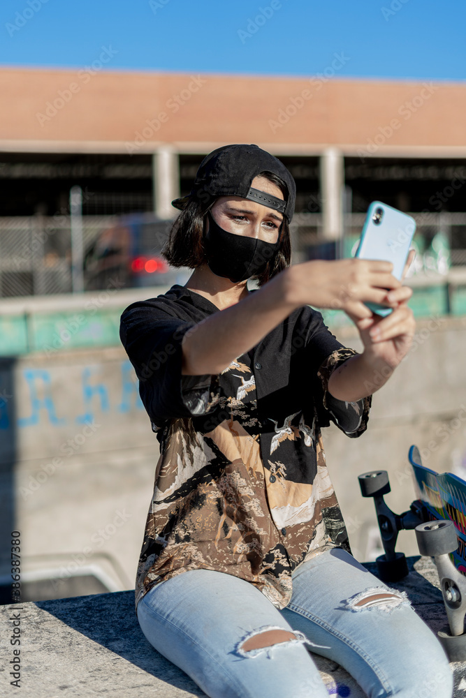 Mujer joven con mascarilla con skate monopatín en skatepark haciendo fotos selfies - obrazy, fototapety, plakaty 