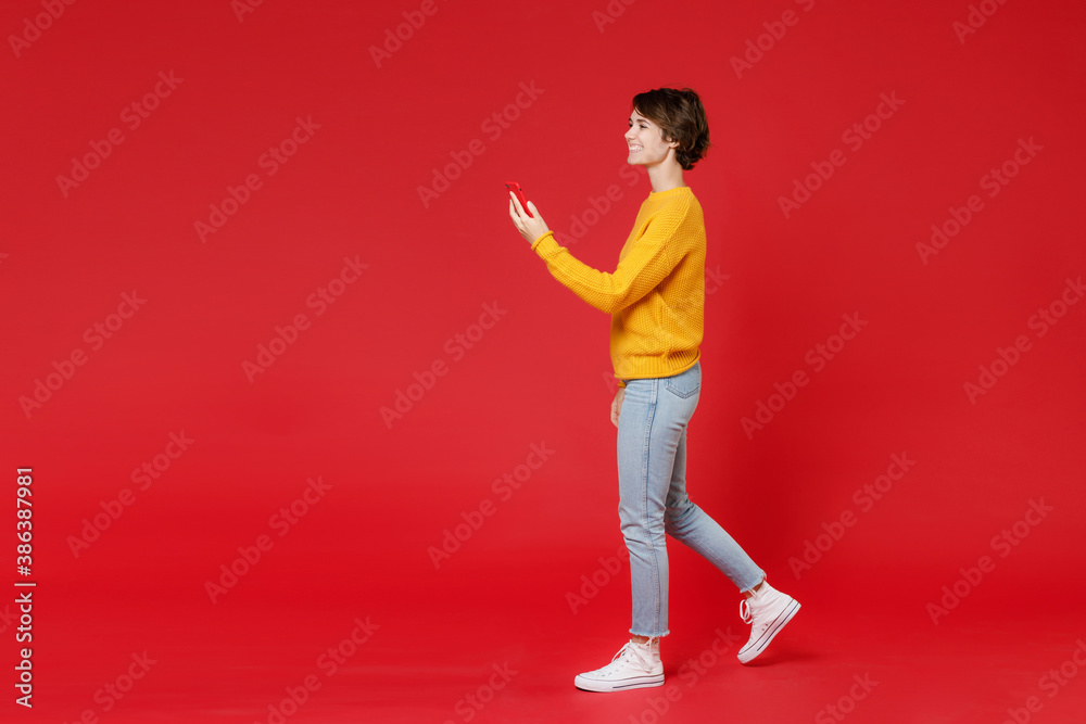 Full length side view of smiling beautiful young brunette woman 20s in basic casual yellow sweater using mobile cell phone typing sms message isolated on bright red colour background studio portrait. Full length side view of smiling beautiful young brunette woman 20s in basic casual yellow sweater using mobile cell phone typing sms message isolated on bright red colour background studio portrait.