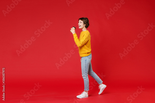 Full length of excited cheerful beautiful young brunette woman 20s wearing casual yellow sweater hold paper cup of coffee or tea looking aside isolated on bright red colour background studio portrait.
