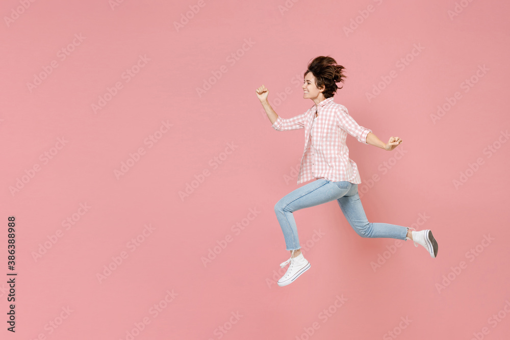 Full length side view of smiling cheerful funny young brunette woman 20s wearing casual checkered shirt jumping like running clenching fists isolated on pastel pink colour background, studio portrait.