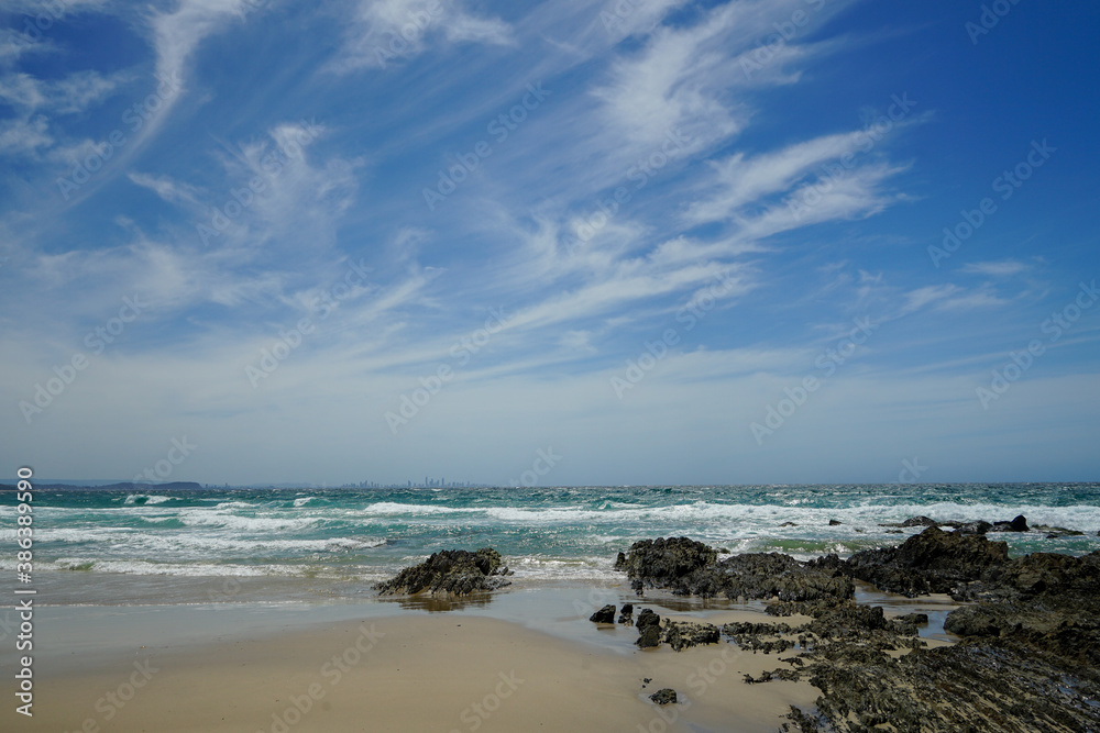 Surf beach on a windy day. View from sandy shore over rocks and choppy sea to city skyline on the horizon, with windswept cirrus clouds against a blue sky.  Coolangatta, Queensland, Australia. 