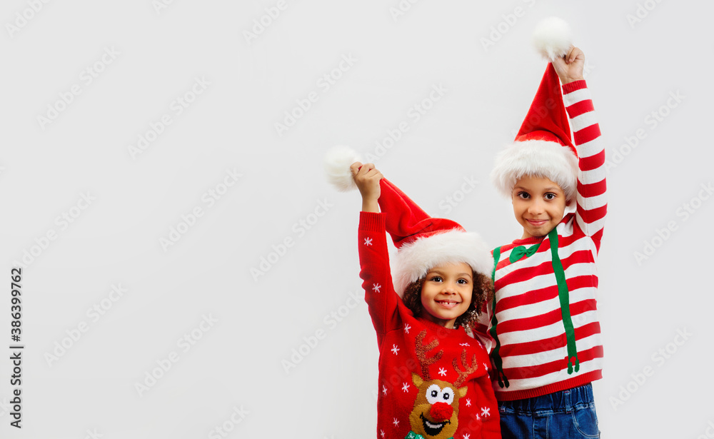 Two happy children in Christmas costumes hold up red Santa hats in Greeting