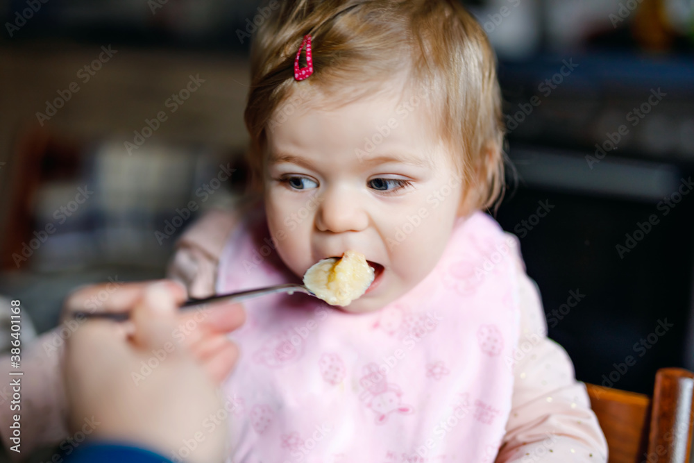 Adorable baby girl eating from spoon mashed vegetables and puree. food, child, feeding and people concept -cute toddler, daughter with spoon sitting in highchair and eating at home. Adorable baby girl eating from spoon mashed vegetables and puree. food, child, feeding and people concept -cute toddler, daughter with spoon sitting in highchair and eating at home.