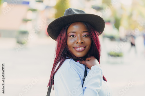 smiling young African American girl with pink hair wearing a hat walking down the street on a sunny day.