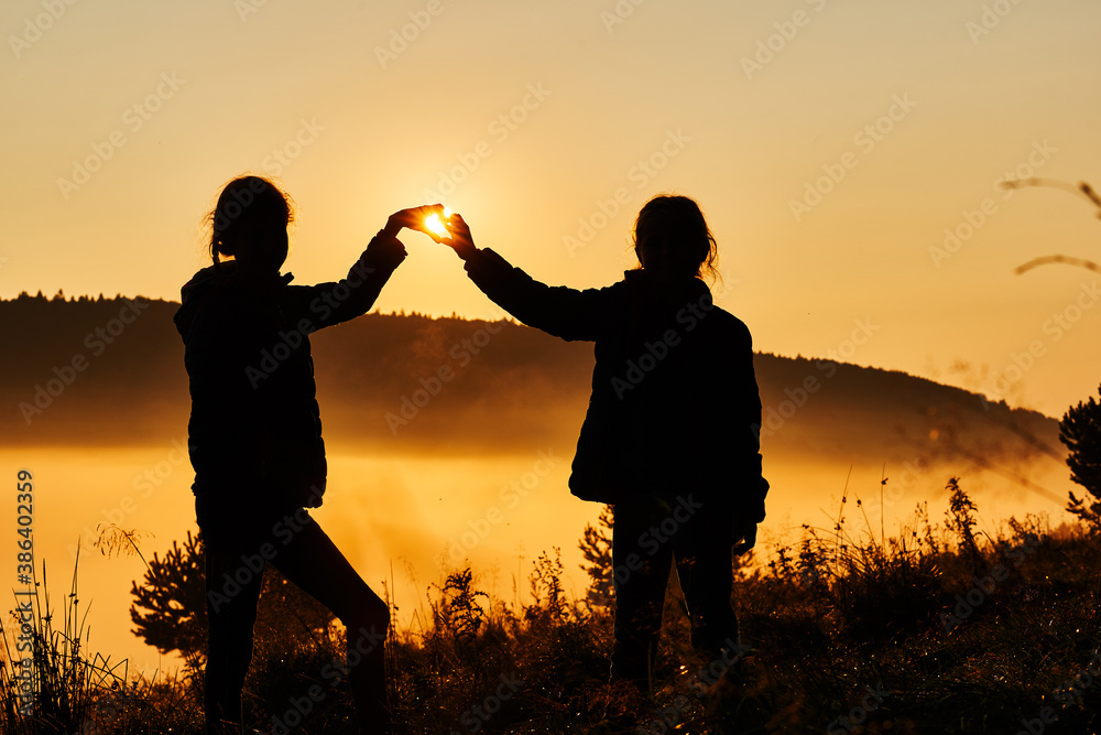 Beautiful fog in the morning. Silhouettes of two girls making heart with their hands against a beautiful golden sunrise.