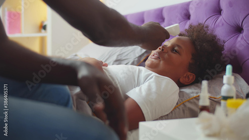 African dad using infrared thermometer to check temperature of little sick boy lying in bed