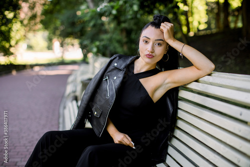 Beautiful sexy Asian woman with ponytail hair style, wearing elegant black overalls and leather jacket, relaxing on bench at urban green summer park. People portraits outdoors © sofiko14