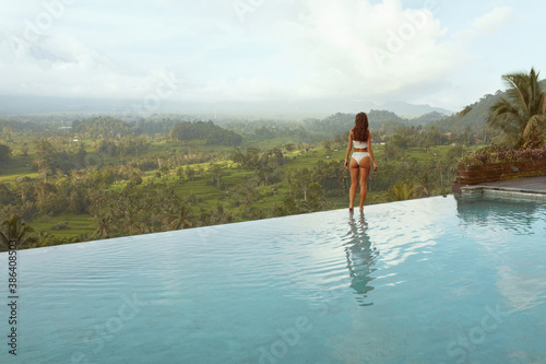 Beautiful Girl   s In Bikini In Infinity Pool Full-Length Portrait. Side View Of Young Woman With Sexy Body In White Swimsuit Posing Against Mount Agung At Tropical Resort In Bali  Indonesia.