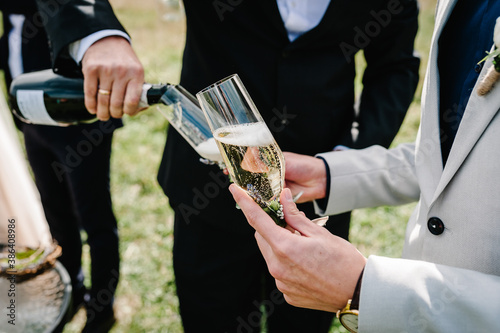 The groom pours champagne into a glass on the background of the friends on nature at the wedding ceremony. Close up. Toast.