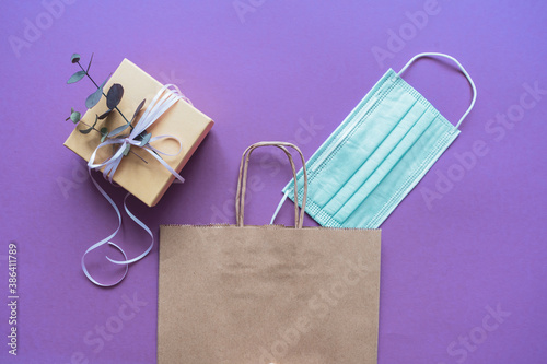 Eine Papier Einkaufstasche mit einer Geschenk Box und einem blauen Mundschutz auf einem lila Hintergrund. Isoliert, Flat lay, shoppen.
