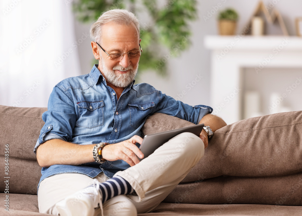 Cheerful aged man using tablet on couch.