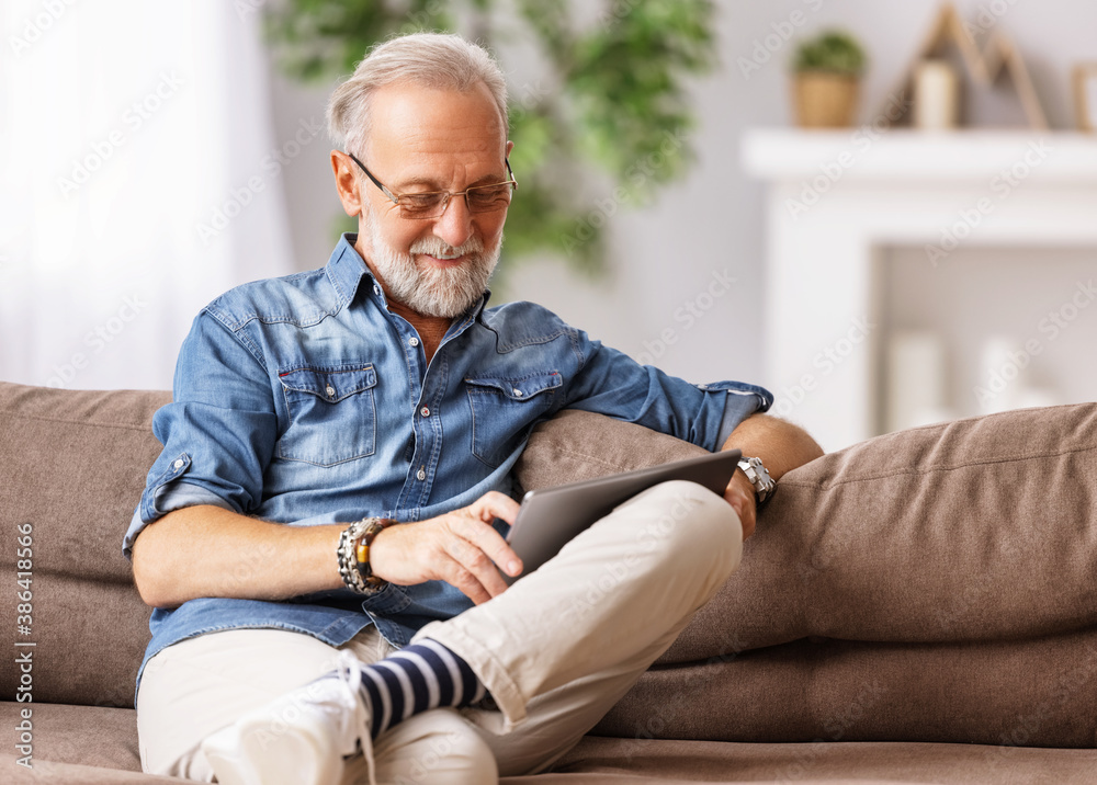 Fototapeta premium Cheerful aged man using tablet on couch.