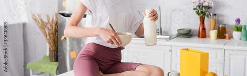 cropped view of young woman holding bottle of milk and bowl in kitchen, panoramic concept