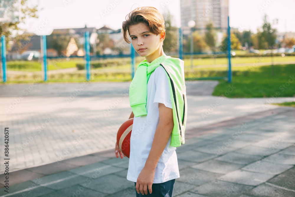 A beautiful teenager holds a basketball in his hands and looks at the camera. Portrait of a boy with a ball in his hands. Sports, education, leisure