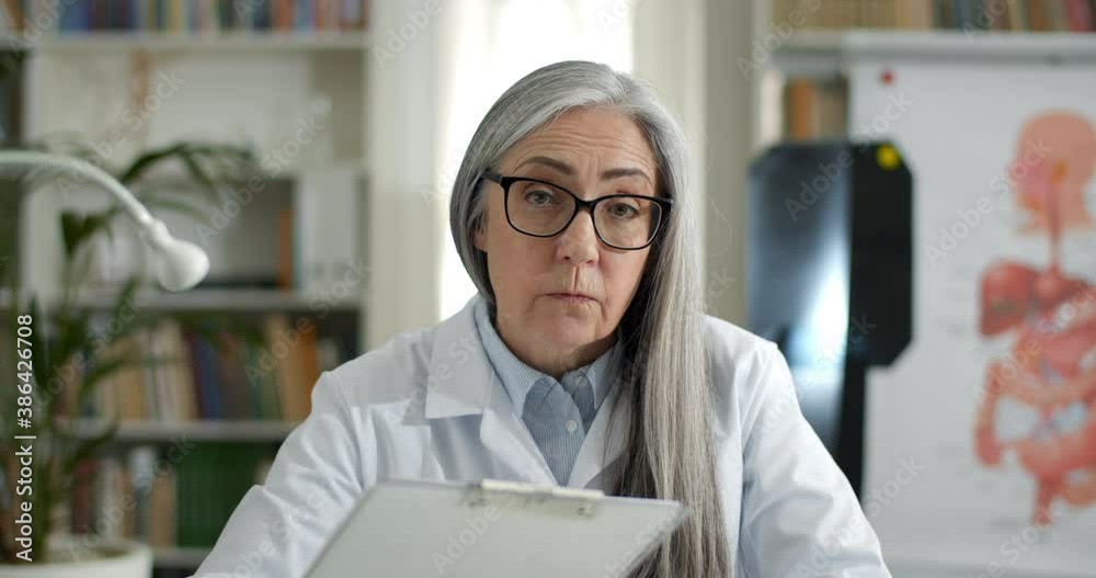 Crop view of female elderly doctor talking with patient and looking to camera while filling up medical form. Woman in white rob holding paper holder and writing while having video call Crop view of female elderly doctor talking with patient and looking to camera while filling up medical form. Woman in white rob holding paper holder and writing while having video call