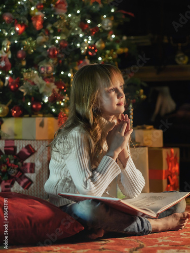 Happy young girl reading a story book in a cozy dark living room on Christmas eve. Celebrating Xmas at home. Christmas tree on the background.