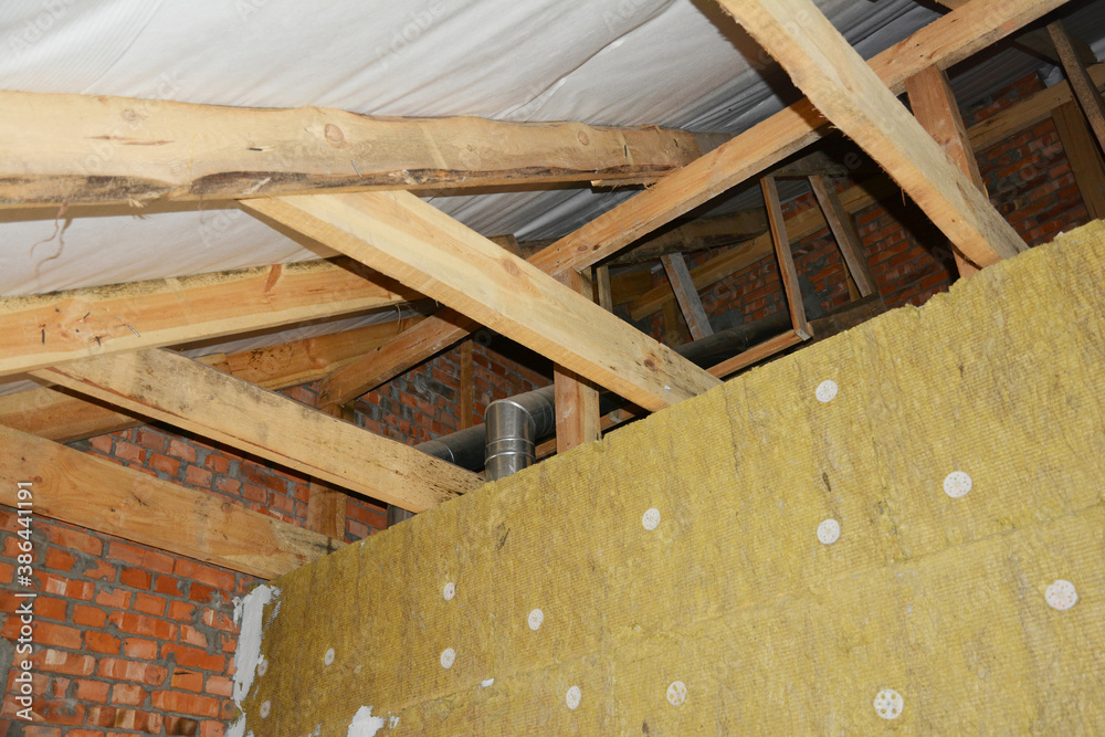 A view from inside a house attic construction on unfinished roof with wooden frame, roof beams, rafters, ceiling joists, trusses, vapor barrier installed and a wall with mineral wool insulation. A view from inside a house attic construction on unfinished roof with wooden frame, roof beams, rafters, ceiling joists, trusses, vapor barrier installed and a wall with mineral wool insulation.