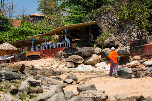 A woman with an orange handkerchief on her shoulders walks along the rocky coast along the village houses in Goa, India