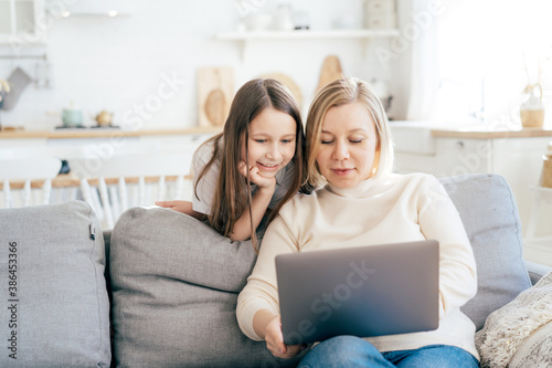 Smiling mom and daughter surf in a laptop on the couch at home. Having fun online, internet education, working from home