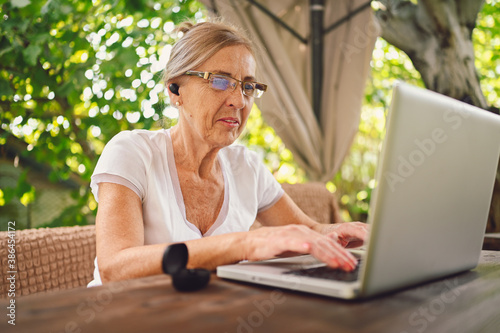 Happy senior woman use wireless headphones working online with laptop computer outdoor in garden © Алина Троева