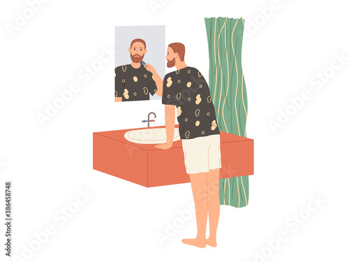 Man shaves his beard with an electric razor while standing in the bathtub by the mirror