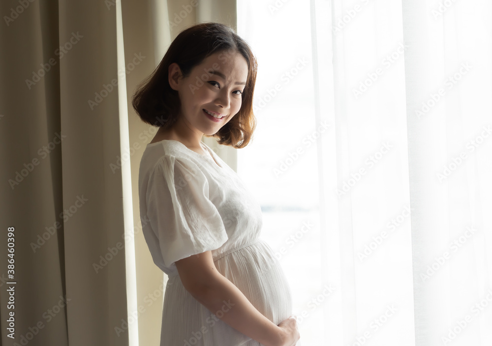 Asian pregnant woman wearing white dress and standing beside window Asian pregnant woman wearing white dress and standing beside window