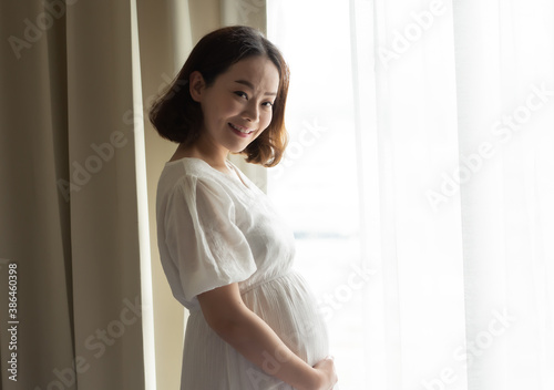 Asian pregnant woman wearing white dress and standing beside window